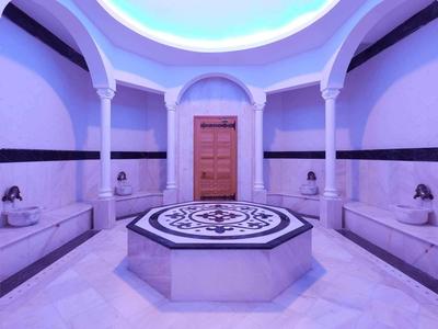 Rundes Hamam mit weißen Marmorbänken, Säulen, blauem Licht und zentralem Wasserspender.