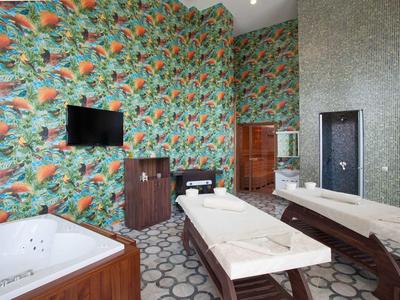 Moderner Wellnessraum mit Massageliege, Whirlpool, buntem Vogel-Tapetenmuster und großem Spiegel.