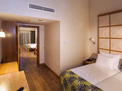 Schlichtes Hotelzimmer mit Einzelbett, Holzboden, Schreibtisch und Blick in angrenzendes Zimmer.