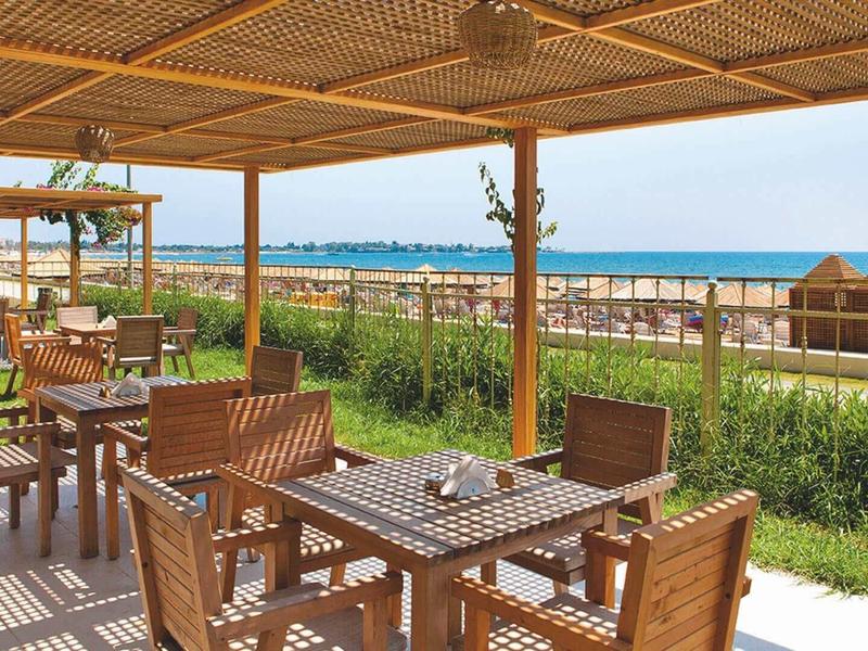 Offenes Café mit Holztischen und Stühlen, Blick aufs Meer, grüner Rasen und Strand im Hintergrund.