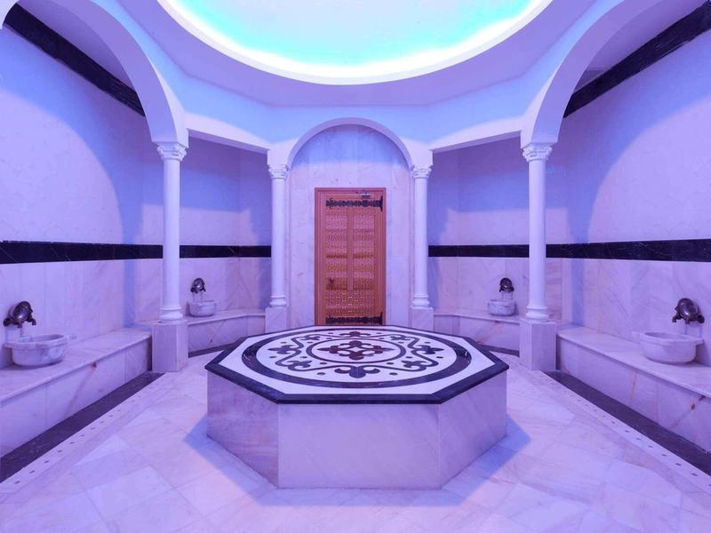 Rundes Hamam mit weißen Marmorbänken, Säulen, blauem Licht und zentralem Wasserspender.