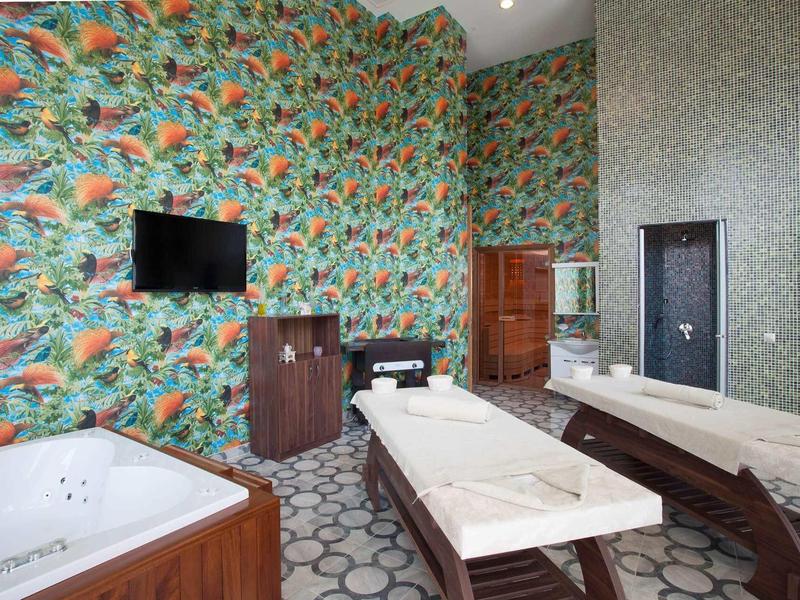 Moderner Wellnessraum mit Massageliege, Whirlpool, buntem Vogel-Tapetenmuster und großem Spiegel.