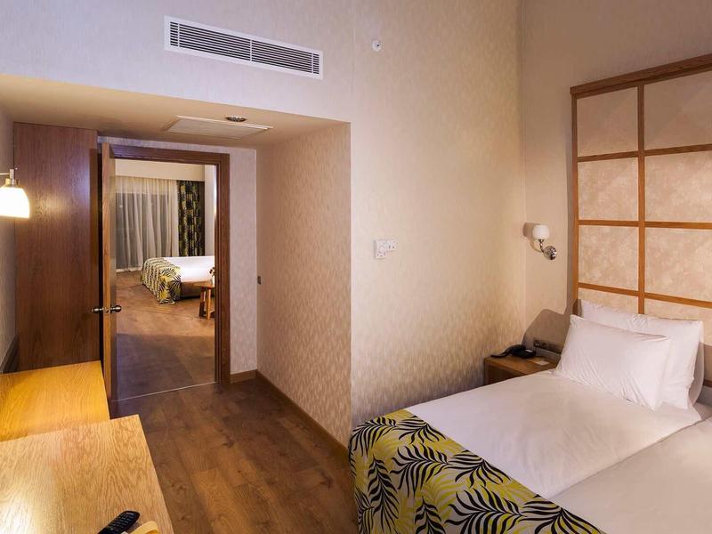 Schlichtes Hotelzimmer mit Einzelbett, Holzboden, Schreibtisch und Blick in angrenzendes Zimmer.