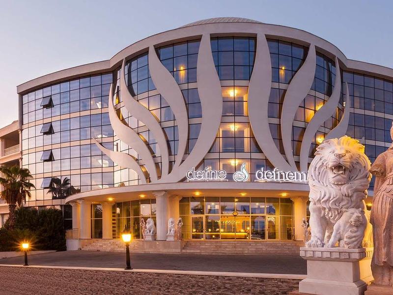 Modernes Glasgebäude mit geschwungener Fassade und zwei steinernen Skulpturen im Vordergrund bei Sonnenuntergang.
