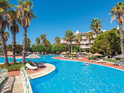 Grande piscina con palme e lettini davanti a un hotel sotto un cielo sereno.