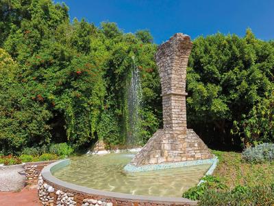 Fontana di pietra con getto alto davanti a una fitta foresta verde e cielo blu.