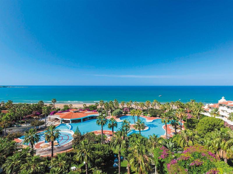 Pohled na hotelový bazén s palmami a mořem v pozadí pod jasnou modrou oblohou.