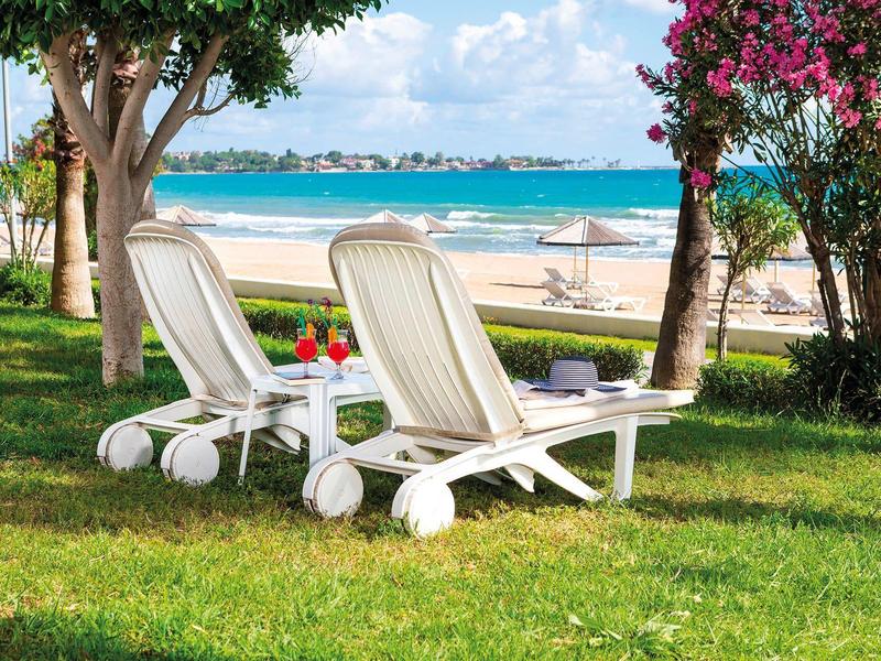 Dvě bílé lehátka na zelené trávě s výhledem na písečnou pláž a moře.