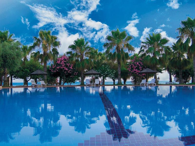 Velký bazén s čistou vodou, obklopený palmami a kvetoucími rostlinami pod modrou oblohou.