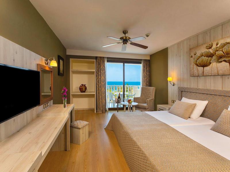 Moderní hotelový pokoj s výhledem na moře, manželskou postelí, posezením a televizí na zdi.