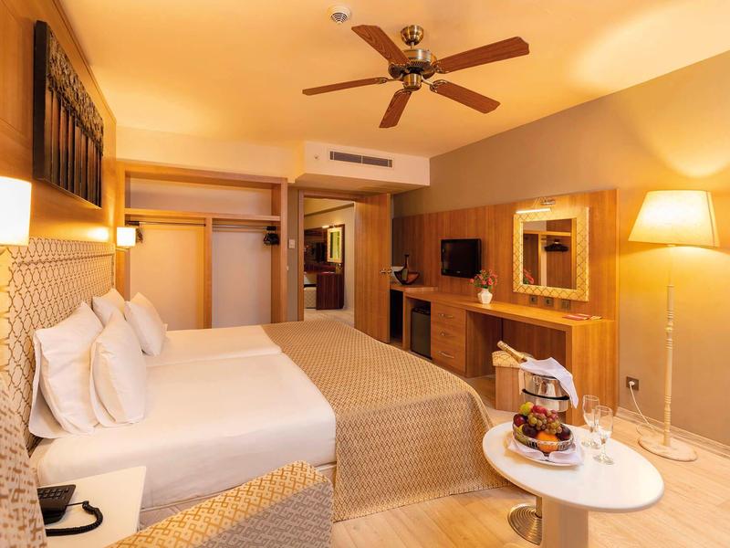 Pohodlný hotelový pokoj s velkou postelí, stropním ventilátorem a stolem s občerstvením.