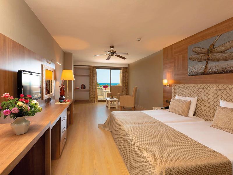 Světý hotelový pokoj s manželskou postelí, psacím stolem, pohovkou a oknem s výhledem na moře.