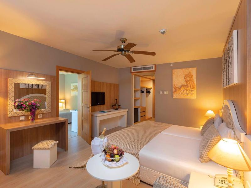 Světly hotelový pokoj s velkou postelí, stropním ventilátorem a útulným osvětlením.