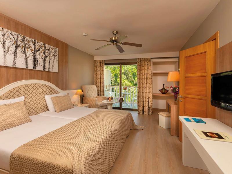 Světý hotelový pokoj s velkou postelí, posezením, televizí a balkonem s výhledem na zahradu.