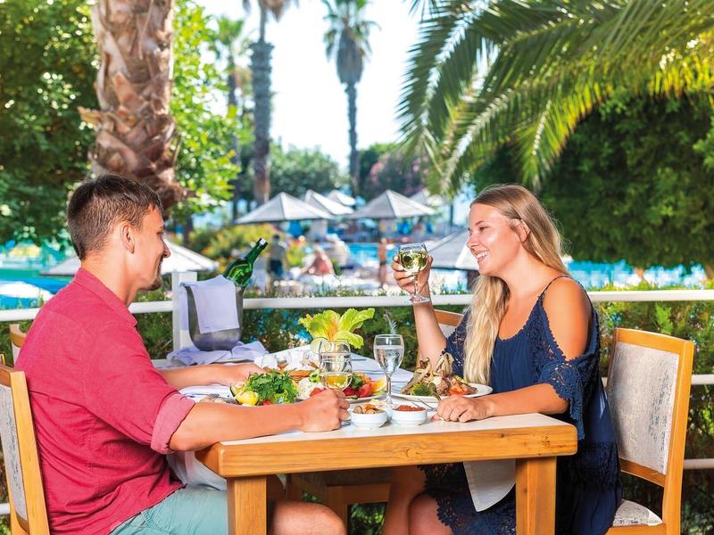 Pár sedí venku u prostřeného stolu, pije víno a vychutnává si jídlo pod palmami.