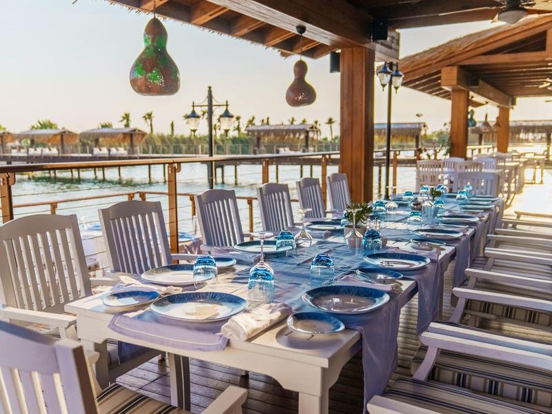 Langer gedeckter Esstisch mit weißen Stühlen auf einer Terrasse mit Blick auf Wasser.