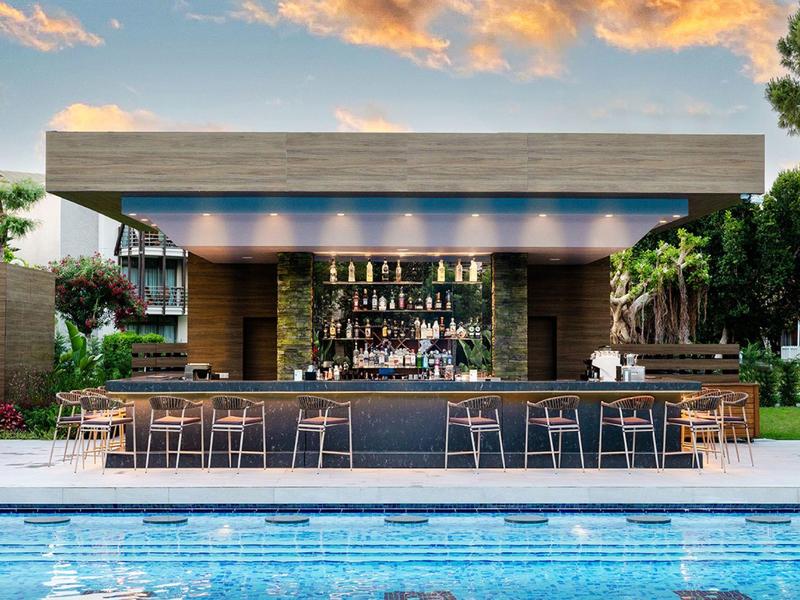 Moderner Poolbar mit Holzstruktur, metallischen Barhockern und blauem Schwimmbad im Vordergrund.