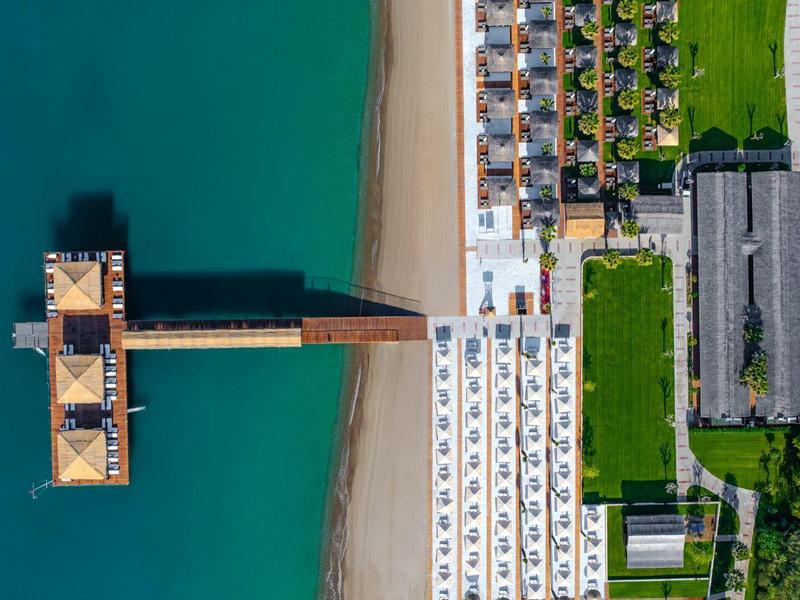Luftaufnahme eines Hotels mit Weinbar auf einem Holzsteg über blauem Wasser und Liegestühlen am Strand.