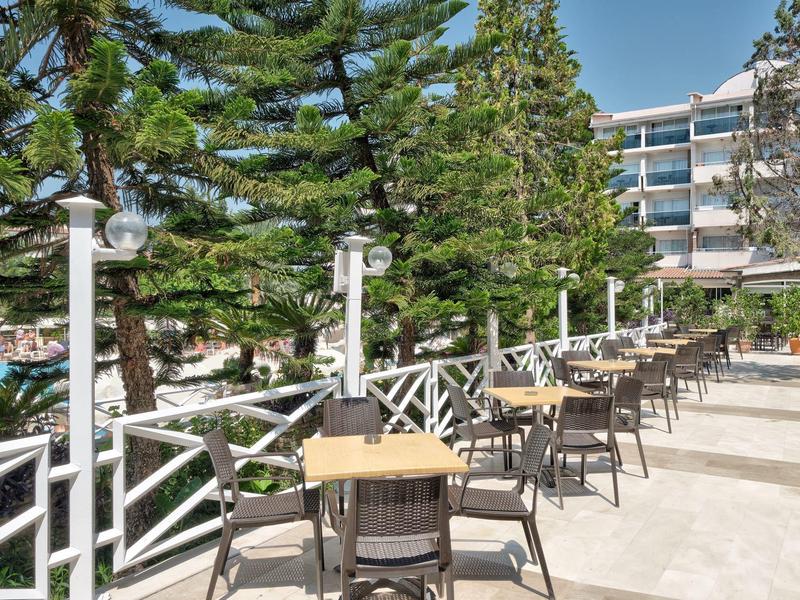 Terrasse d'hôtel avec plusieurs tables et chaises à côté de grands arbres et d'un bâtiment à plusieurs étages.