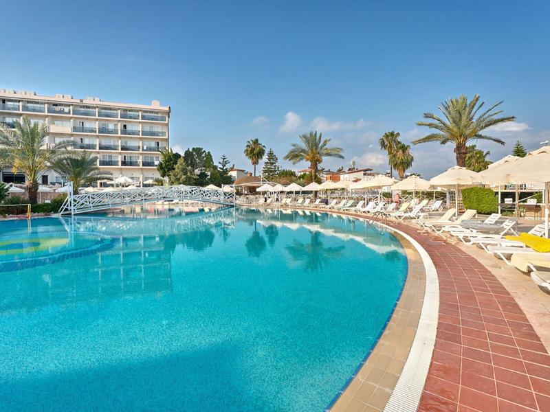 Grande piscine d'hôtel avec chaises longues, parasols et palmiers sous un ciel clair.