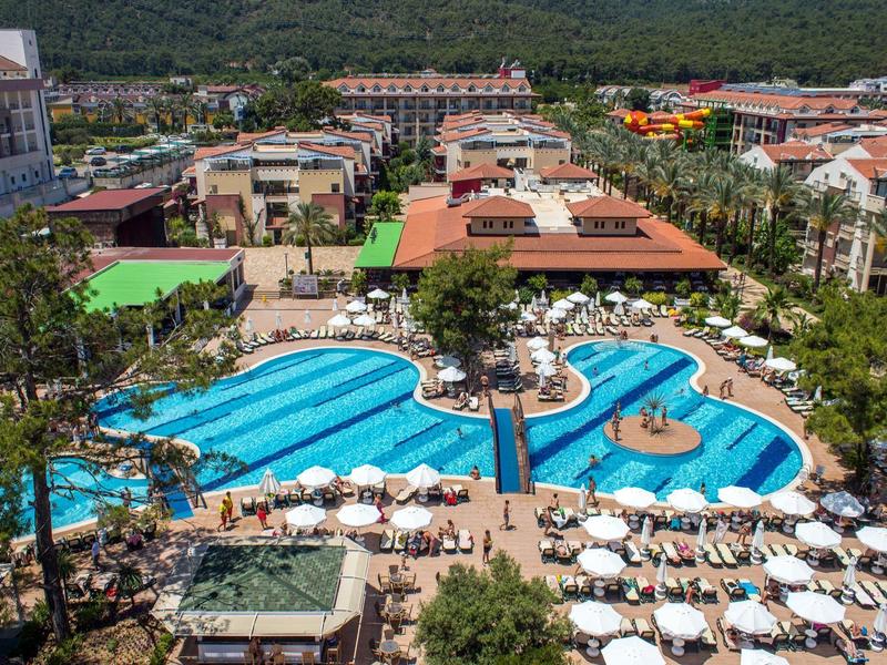Grand piscine d'hôtel avec parasols et chaises longues, entourée de bâtiments et de montagnes vertes.
