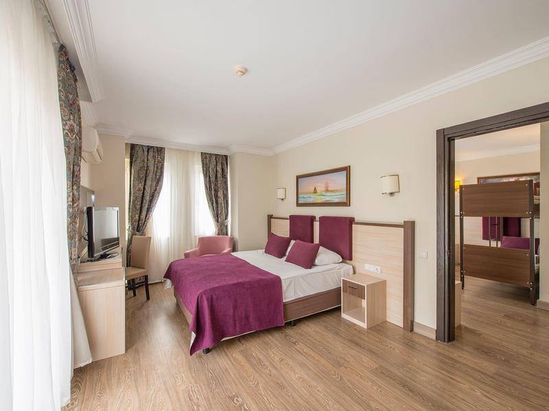 Chambre d'hôtel lumineuse avec lit double, couvre-lit violet, chaise et bureau à côté d'une grande fenêtre.