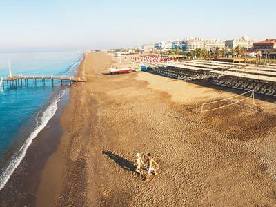 Široká písečná pláž s klidným mořem, molem a budovami na pozadí za jasného dne.