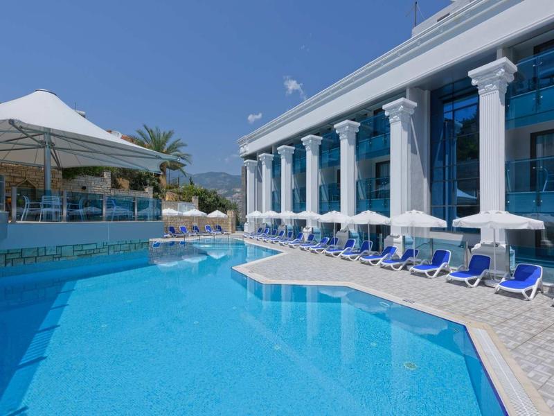 Piscina esterna moderna con lettini blu e ombrelloni accanto a un hotel con colonne.