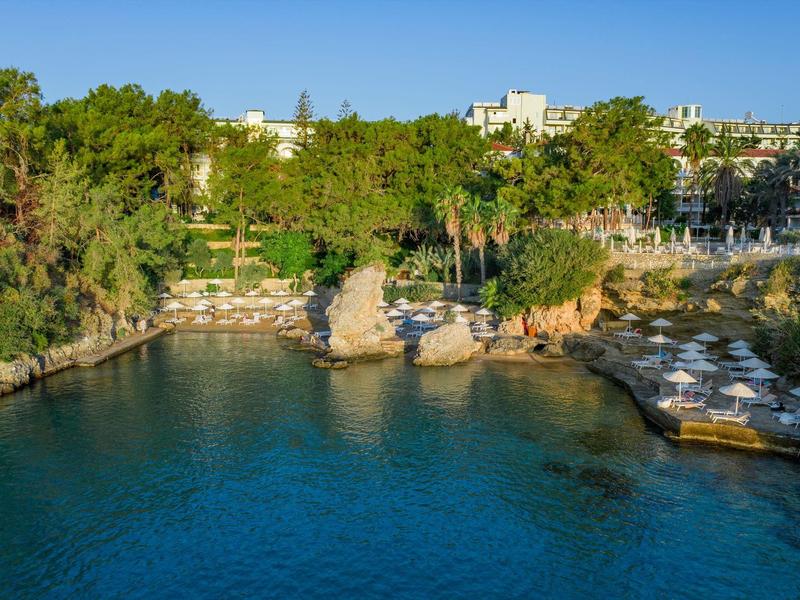 Klares Meer mit felsiger Küste und grünen Bäumen vor weißen Gebäuden bei sonnigem Himmel.