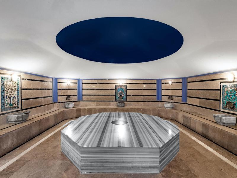 Moderner Raum mit hölzernen Sitzbänken, Symmetrie, Metallobjekt in der Mitte und blauer runder Deckenöffnung.