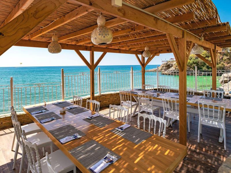 Holzterrasse mit Tischen und Stühlen am Meer, überdacht mit Bambusdach und Blick auf klares Wasser.