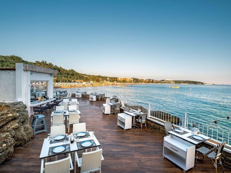 Offenes Restaurant mit weißen Möbeln auf Holzdeck am Meer unter klarem Himmel.