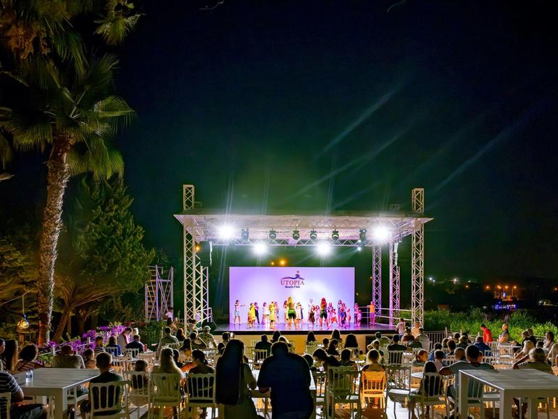 Freiluftbühne mit Aufführung bei Nacht, Publikum an Tischen, umgeben von Bäumen und dunklem Himmel.