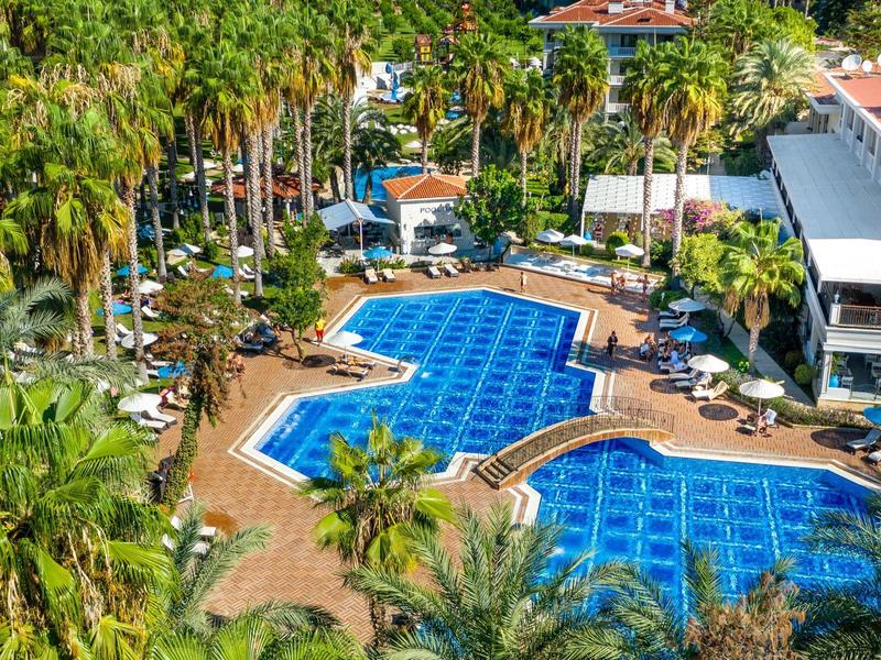 Großer Hotelpool mit blauen Fliesen, umgeben von Palmen, Liegestühlen und einem weißen Gebäude.