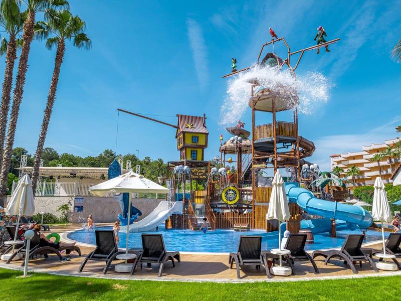 Wasserpark mit Rutschen, Palmen, Liegestühlen und einem großen Spielschiff unter blauem Himmel.