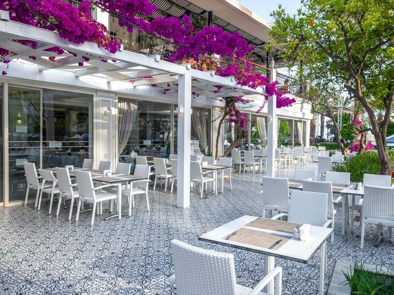 Freiluftrestaurant mit weißen Tischen und Stühlen, überdacht mit blühenden Bougainvillea-Pflanzen.