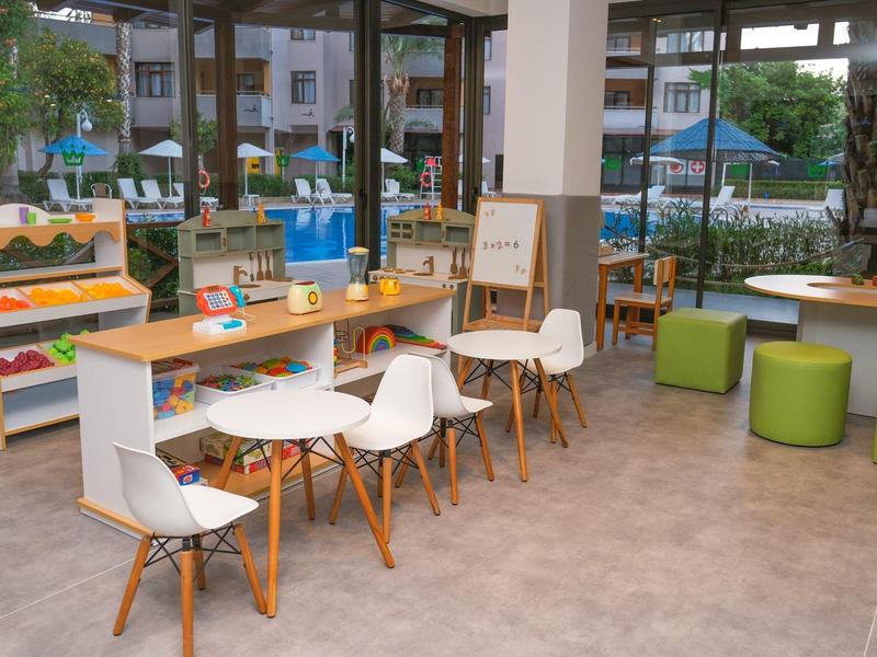 Hell gestalteter Lernraum mit Holzstühlen, weißen Tischen und grünen Sitzwürfeln vor großen Fenstern.