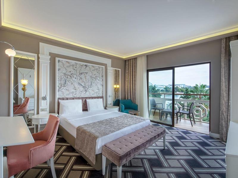 Helles Hotelzimmer mit großem Bett, Sitzbank, Schreibtisch, Balkon und modernem Bodenmuster.