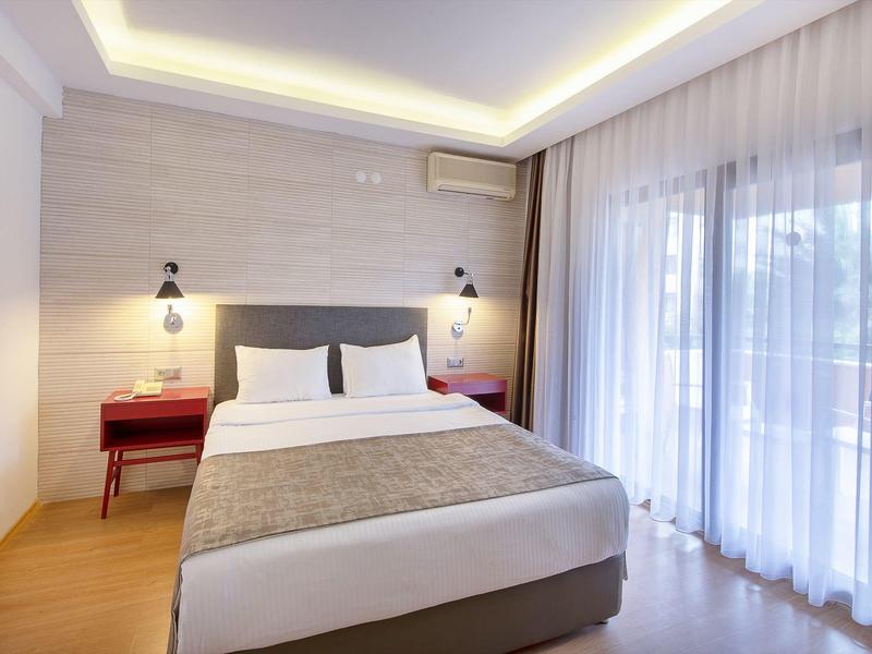 Hell eingerichtetes Schlafzimmer mit Doppelbett, roten Nachttischen und großen Fenstern mit Vorhängen.