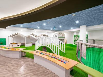 Moderner, heller Spielplatz mit grünem Kunstrasen, weißen Holzbrücken und bunten Bänken.