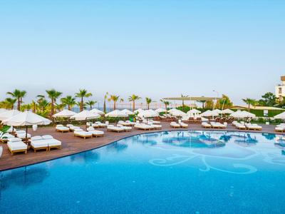 Area piscina dell'hotel con ombrelloni, sdraio e palme sotto un cielo limpido.