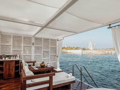 Offene Holzterrasse mit gemütlichem Sofa, Blick auf Meer und Hafen mit Segelboot bei klarem Himmel.