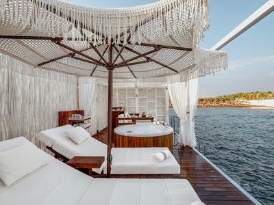 Luxuriöse Terrasse mit weißen Liegen, Sonnenschirm und Holzbadewanne am Meer unter blauem Himmel.
