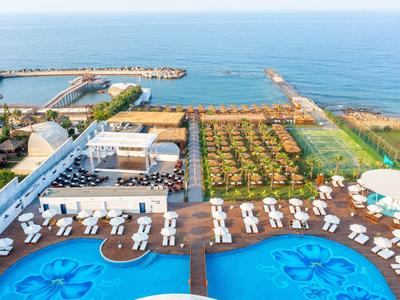 Großer Pool mit Liegestühlen am Meer, dahinter Grünflächen und ruhiges blaues Wasser.