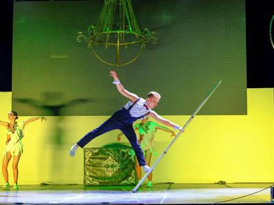 Uno spettacolo colorato sul palco con ballerini davanti a uno sfondo giallo e una grande lampada verde.
