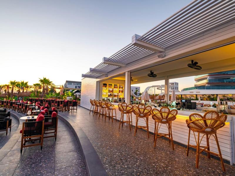 Offene Bar mit Holzstühlen, modernem Design und Blick auf eine Palmenlinie bei Sonnenuntergang.