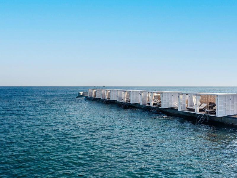 Blaue See mit klarem Himmel und modernem Steg mit weißen Sitzbereichen auf der rechten Seite.