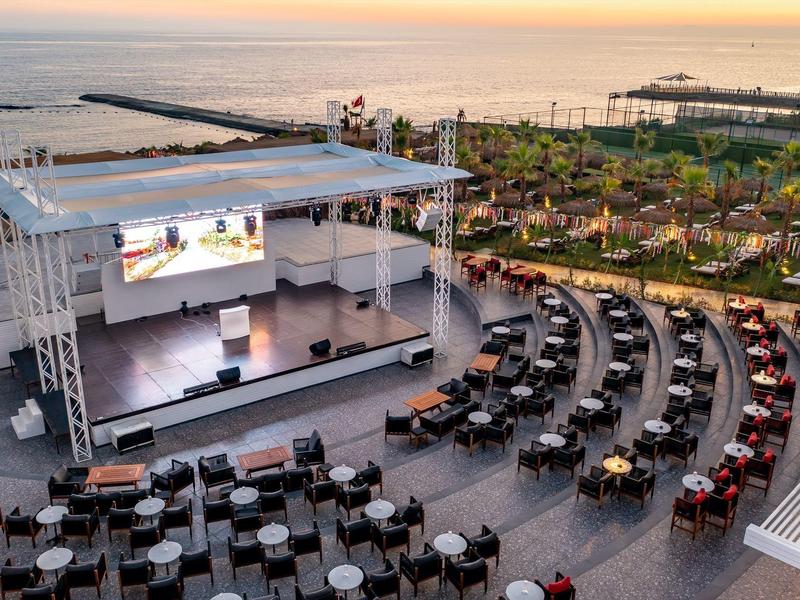 Freiluftbühne mit Halbrund-Sitzreihen und Tischen am Meer bei Sonnenuntergang.