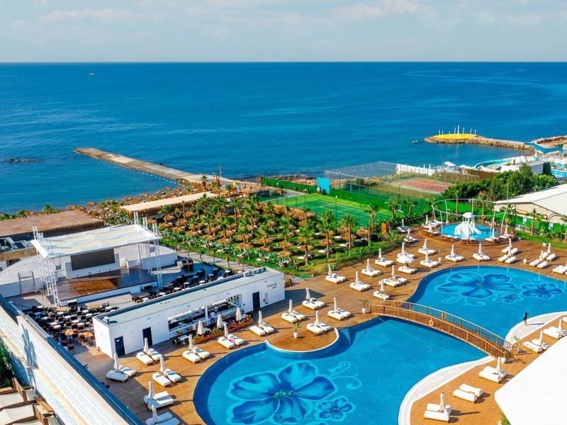 Blick auf ein Hotel mit großen blauen Pools, weißen Sonnenliegen und grünem Garten am Meer.