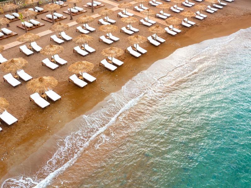 Langer Sandstrand mit Reihen weißer Liegen und sanften Wellen am türkisblauen Wasser.